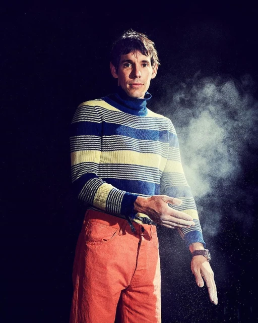 Full studio shot of an adult male wearing a striped turtleneck and orange pants, with white chalk on hands against a black backdrop.