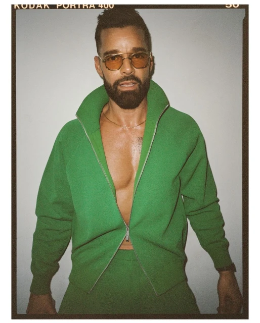 Portrait of adult male with a beard in a green tracksuit and amber glasses posing against a neutral studio backdrop. 
