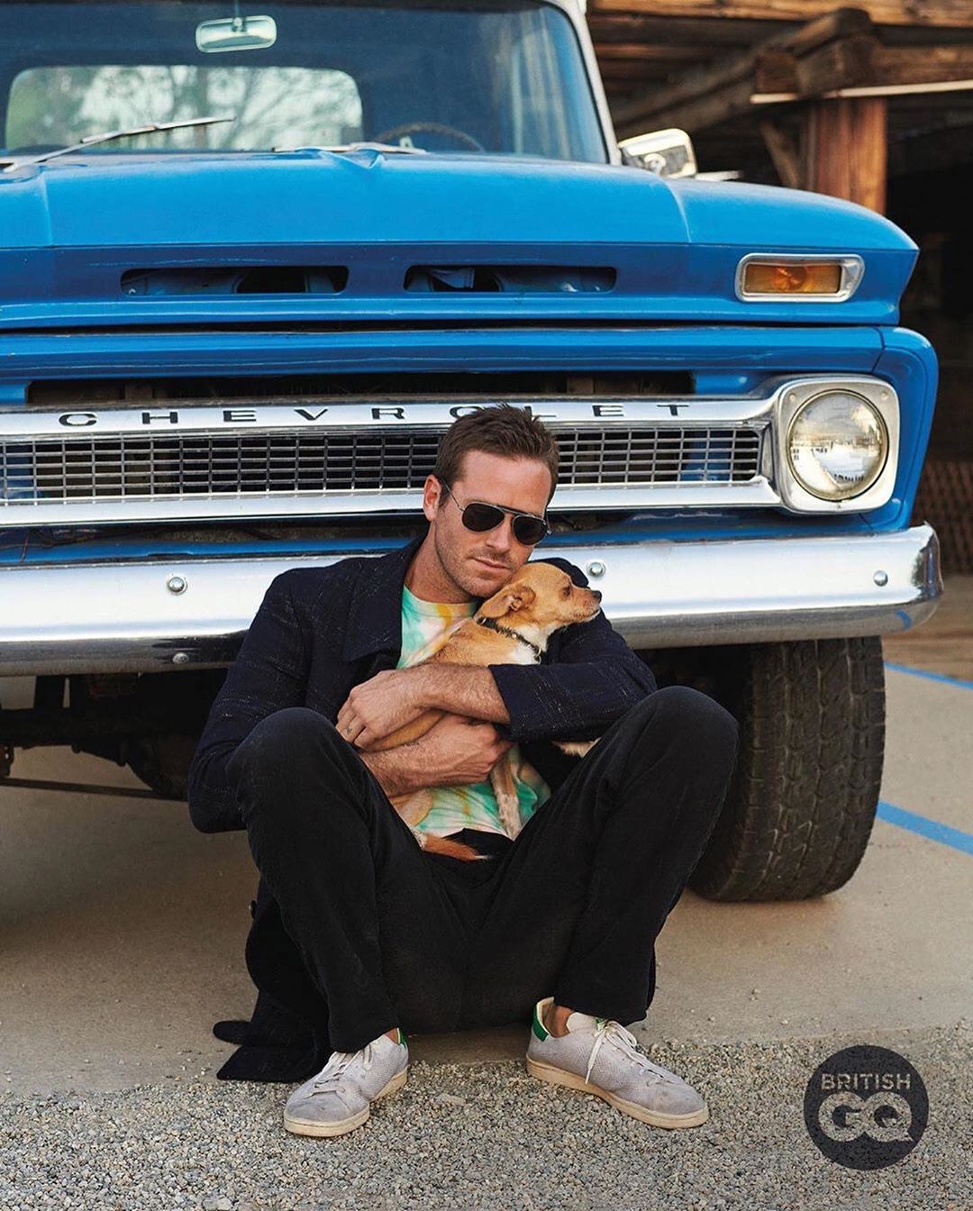 Armie Hammer sitting outdoors with a dog in front of an old blue Chevrolet pickup truck. He wears sunglasses and holds the small dog.