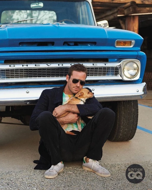 Armie Hammer sitting outdoors with a dog in front of an old blue Chevrolet pickup truck. He wears sunglasses and holds the small dog.