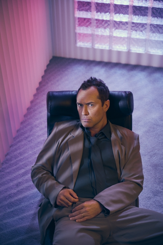 Portrait of an adult male in a suit sitting in a black chair indoors with magenta and purple lighting, creating a surreal atmosphere.