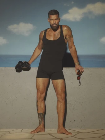 Ricky Martin in a black tank swimsuit holding shoes and sunglasses against a painted sky backdrop