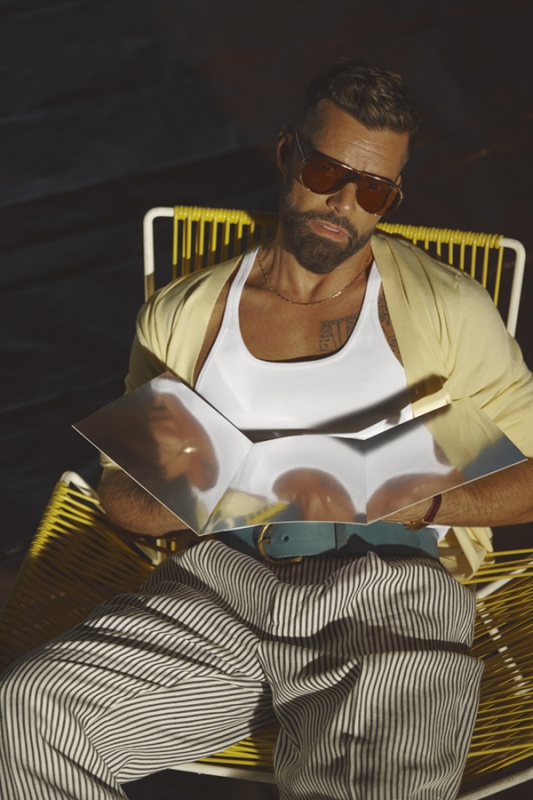 Man in sunglasses relaxes in a yellow chair outdoors, holding a reflective object, wearing striped pants and a white tank top in sunlight.