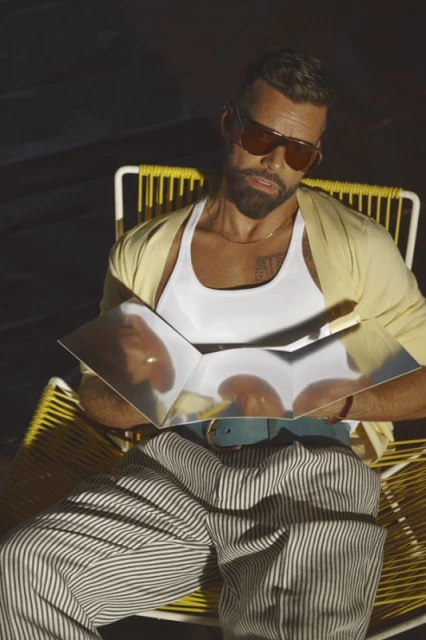 Man in sunglasses relaxes in a yellow chair outdoors, holding a reflective object, wearing striped pants and a white tank top in sunlight.