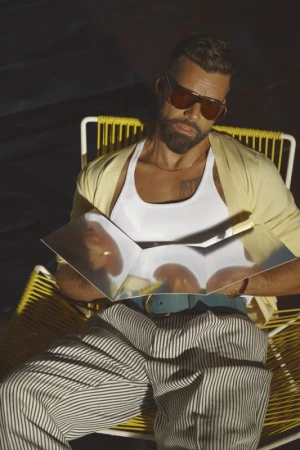 Man in sunglasses relaxes in a yellow chair outdoors, holding a reflective object, wearing striped pants and a white tank top in sunlight.