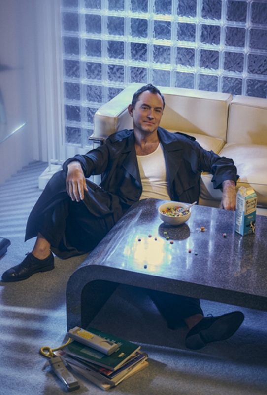 Adult male in a dark coat sits at a coffee table with a bowl of cereal, books, and a milk carton in a casual indoor setting.