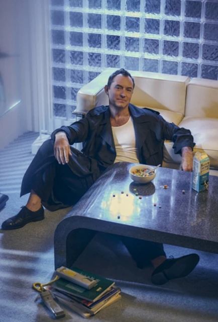 Adult male in a dark coat sits at a coffee table with a bowl of cereal, books, and a milk carton in a casual indoor setting.