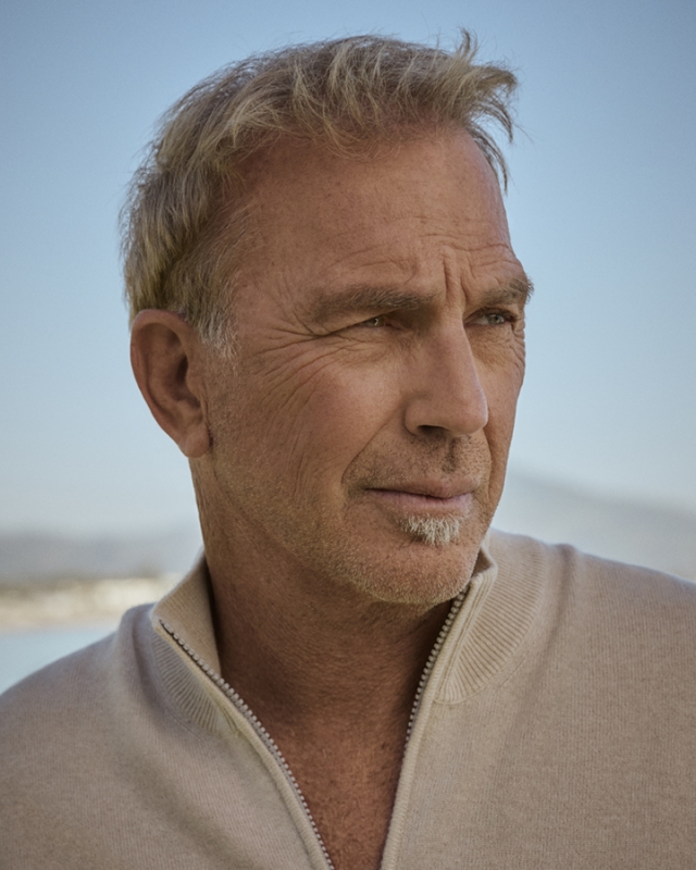 Portrait of adult male with graying blonde hair and light-beige zip-up sweater, set against a blurred blue sky and distant shoreline.