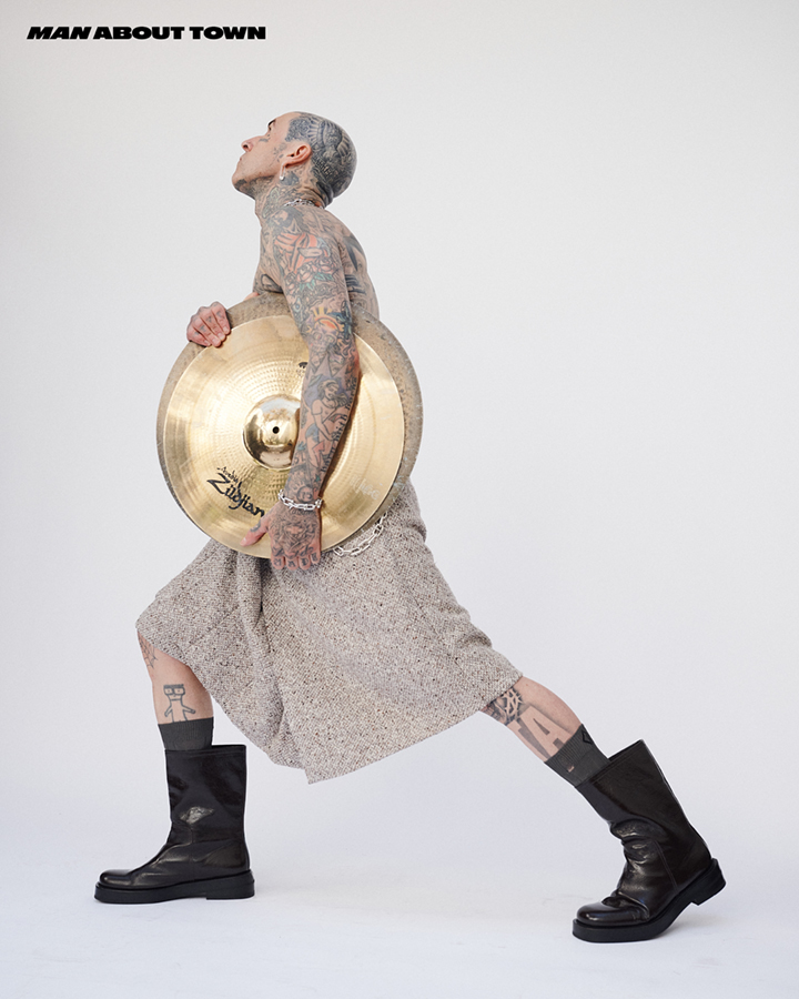 Tattooed adult male poses in a white studio, wearing a skirt and boots while holding cymbals, showcasing a unique blend of fashion and music.