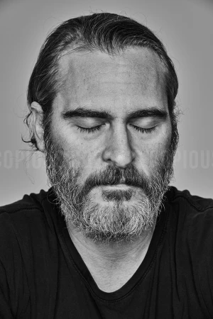 Close-up monochrome portrait of Joaquin Phoenix with eyes closed and full graying beard, wearing a dark t-shirt against a plain backdrop.