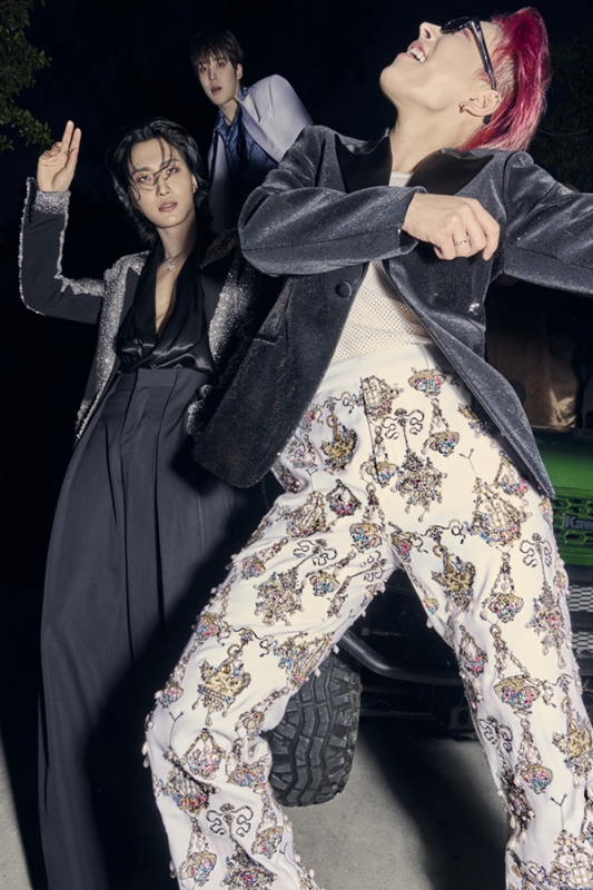 Three Asian adult males pose in formal wear against a dark background, with one man in patterned pants and a dark jacket dominating the frame.