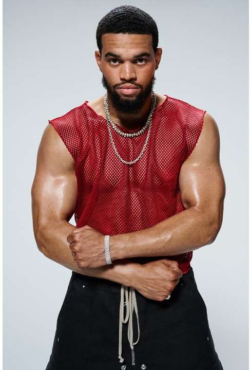 Masculine-presenting person poses indoors wearing a red mesh top and black bottoms, accessorized with jewelry against a light background.