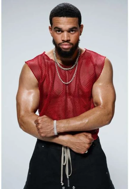 Masculine-presenting person poses indoors wearing a red mesh top and black bottoms, accessorized with jewelry against a light background.