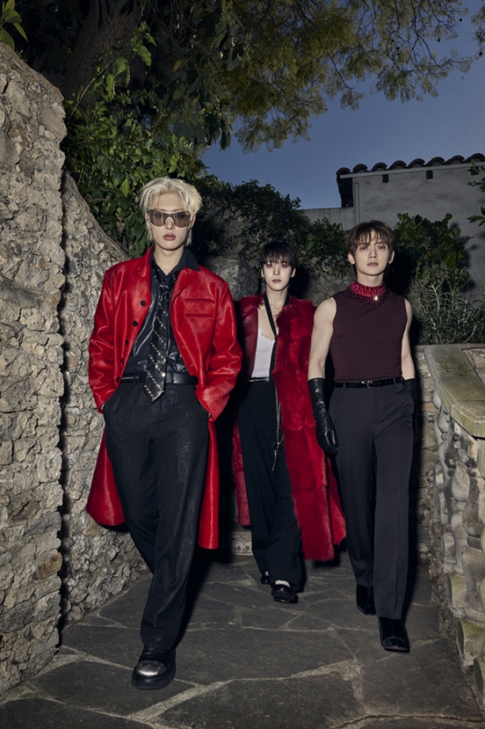 Three stylish adults wearing red jackets and dark pants stand on a stone walkway surrounded by vegetation, showcasing modern fashion.
