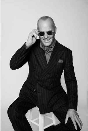Black and white portrait of a smiling adult male wearing sunglasses and a pinstripe suit, seated on a white stool in a photo studio.