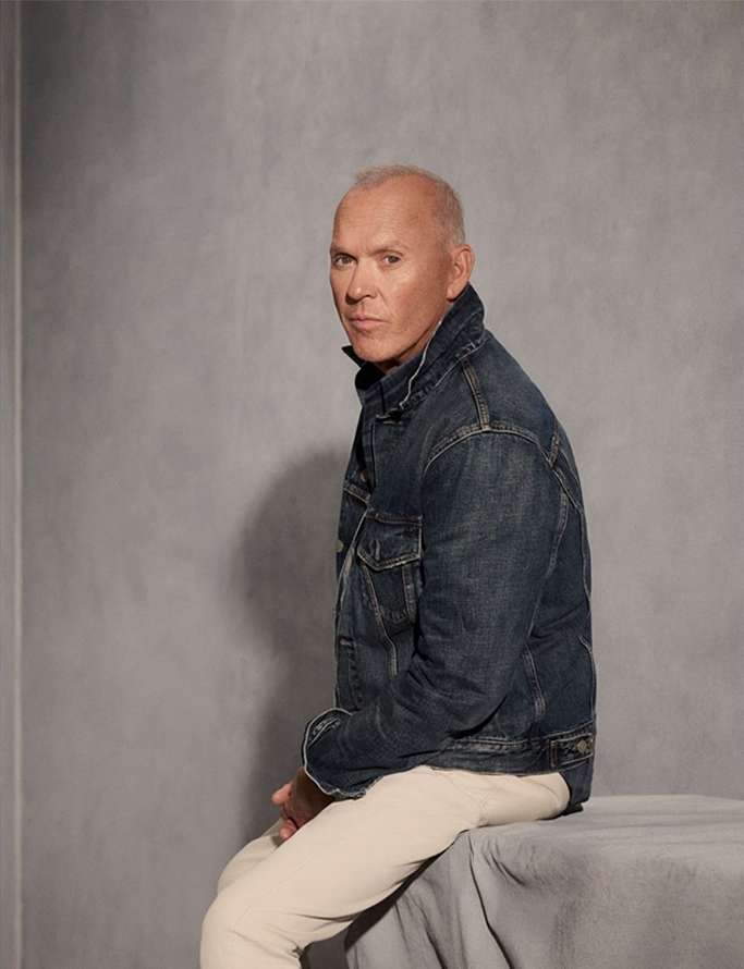 Portrait of actor Michael Keaton in a denim jacket and light trousers, seated against a textured grey wall backdrop in a photo studio.