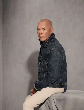Michael Keaton seated in a denim jacket and light trousers against a textured grey studio background photographed by Eric Ray Davidson
