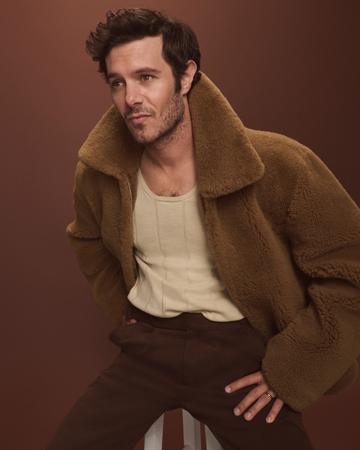 Stylish man in brown jacket poses against a brown background, seated indoors with neutral expression, creating a fashion-forward portrait.