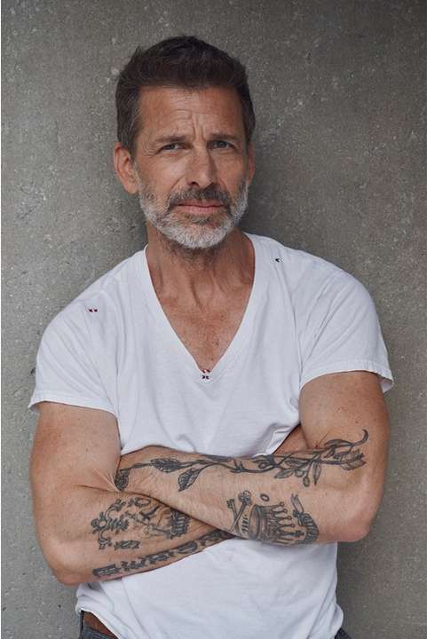 Portrait of a mature man with a light beard and tattoos on his arms posing indoors against a grey wall, wearing a white t-shirt.