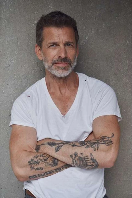 Portrait of a mature man with a light beard and tattoos on his arms posing indoors against a grey wall, wearing a white t-shirt.