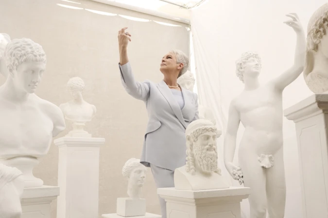 Jamie Lee Curtis poses in a light blue suit surrounded by several white classical Greek-style statue busts and full figure statues.