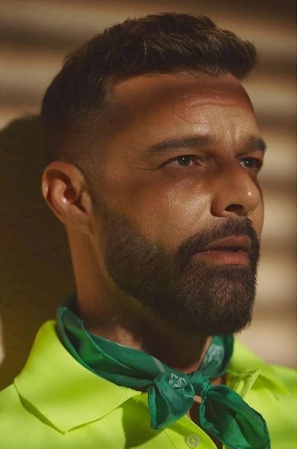 Close-up of a man with short brown hair and a beard, wearing a green scarf and a neon yellow collared shirt against a neutral background.
