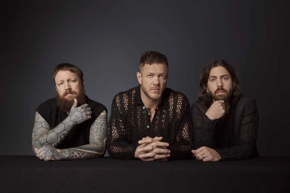 Three bearded musicians pose in a portrait. Dan Reynolds from Imagine Dragons is centered between two bandmates in a dimly lit photo studio.