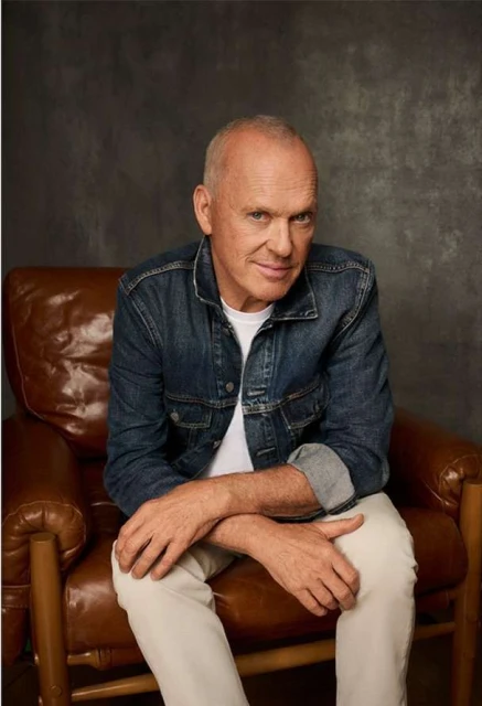 Portrait of a bald adult man in a denim jacket sitting in a leather chair against a mottled brown studio backdrop.