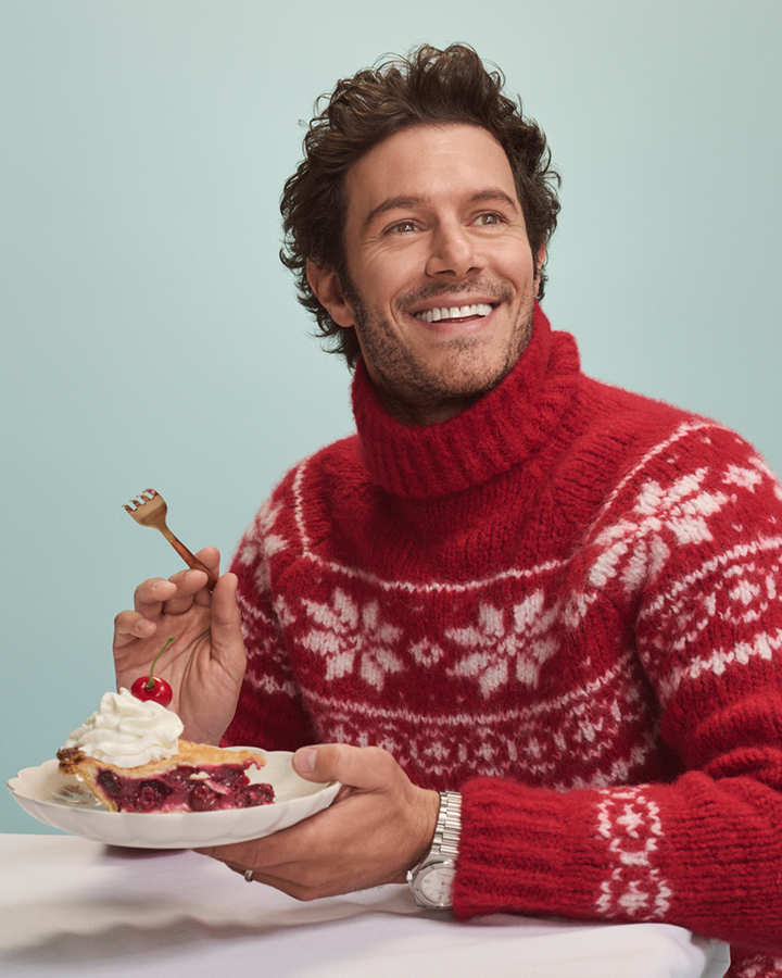Smiling man in red patterned sweater holds a plate of cherry pie topped with whipped cream and a cherry; light blue background.