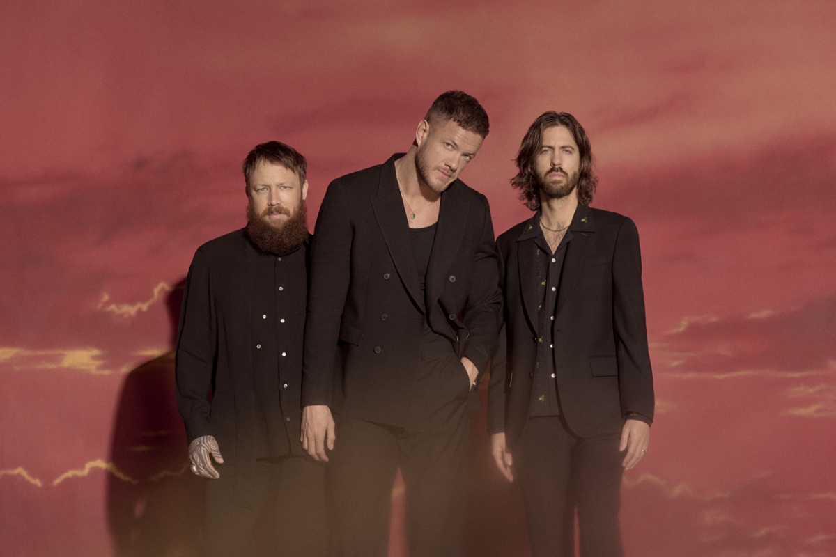 A group studio portrait of three adult male band members in black suits posing against a soft gradient background of pink and orange clouds.