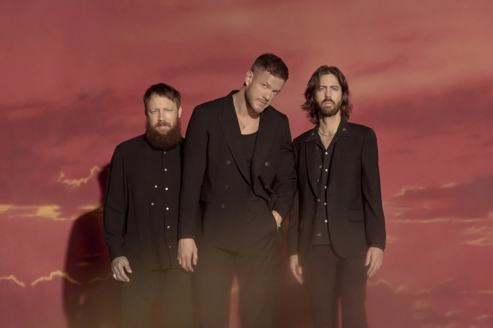 A group studio portrait of three adult male band members in black suits posing against a soft gradient background of pink and orange clouds.