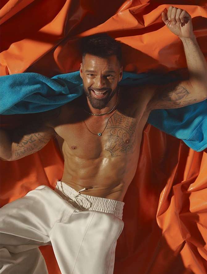 Shirtless adult male smiles while lying on orange fabric with a blue towel.  His arms are up, and he's wearing white pants.