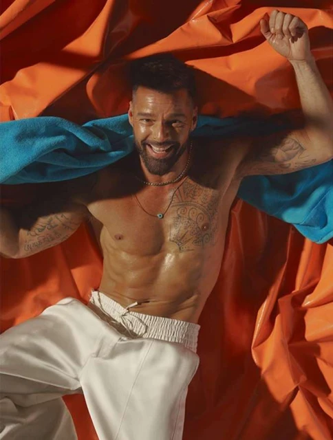 Shirtless adult male smiles while lying on orange fabric with a blue towel.  His arms are up, and he's wearing white pants.