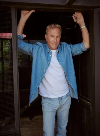 Adult male in denim shirt and jeans stands in doorway, looking at the camera, outside in natural daylight with building in background.