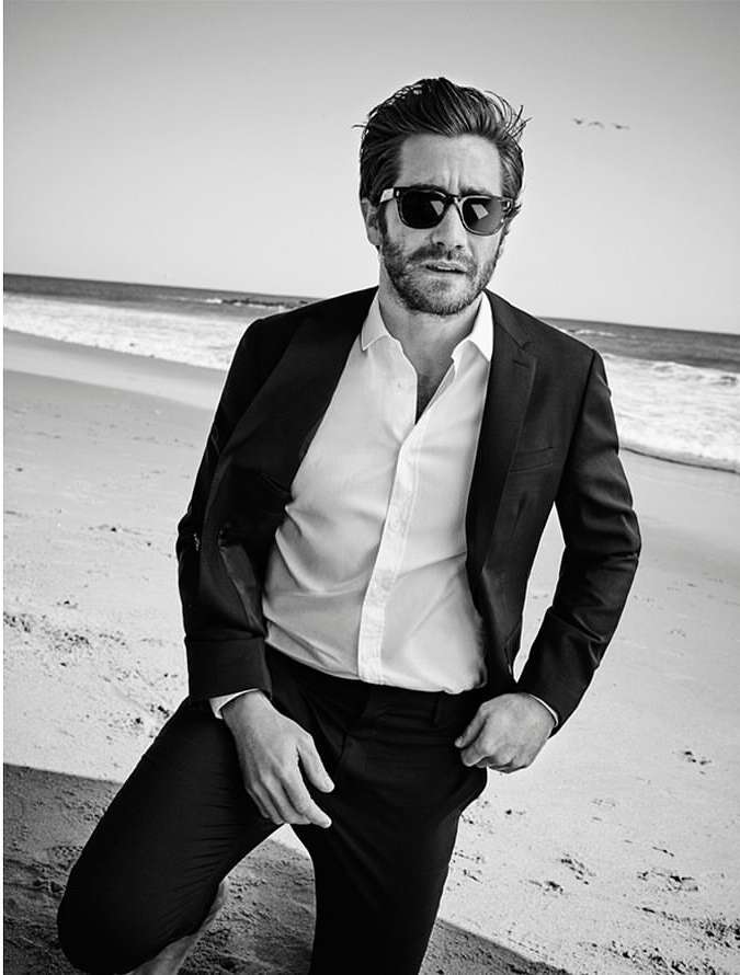 Jake Gyllenhaal poses on a beach in a dark suit and sunglasses. The image is black and white, capturing a sunny day by the ocean.