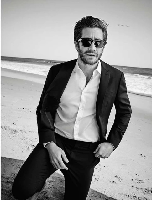 Jake Gyllenhaal poses on a beach in a dark suit and sunglasses. The image is black and white, capturing a sunny day by the ocean.