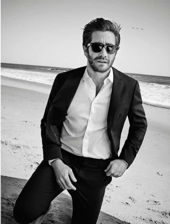Jake Gyllenhaal poses on a beach in a dark suit and sunglasses. The image is black and white, capturing a sunny day by the ocean.