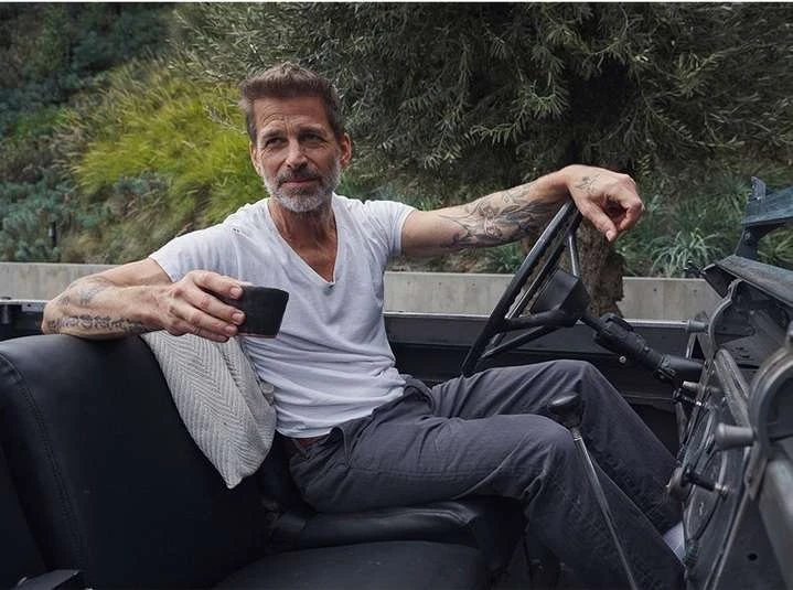 Mature adult man sitting in a convertible car with a cup, wearing a white t-shirt and grey pants in an outdoor setting.