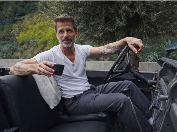 Zack Snyder holding a dark cup while seated in the driver seat of a vintage open-top vehicle wearing a white t-shirt and grey trousers