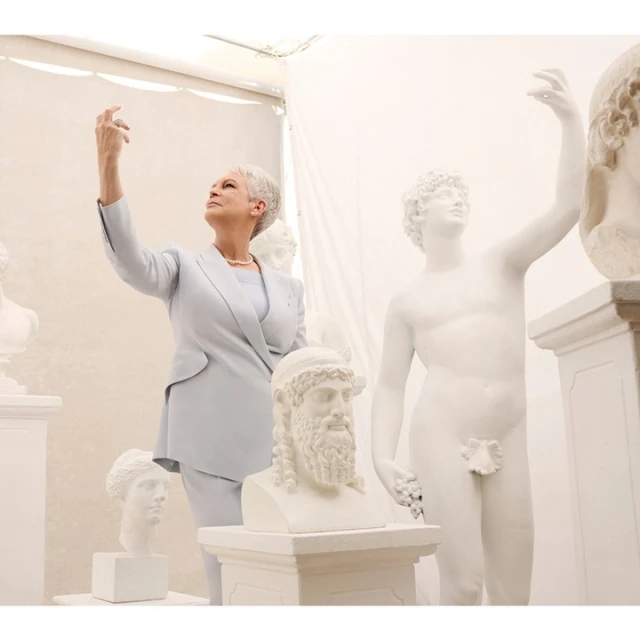 Jamie Lee Curtis poses in a light blue suit alongside various white classical sculpture busts in a bright indoor environment.