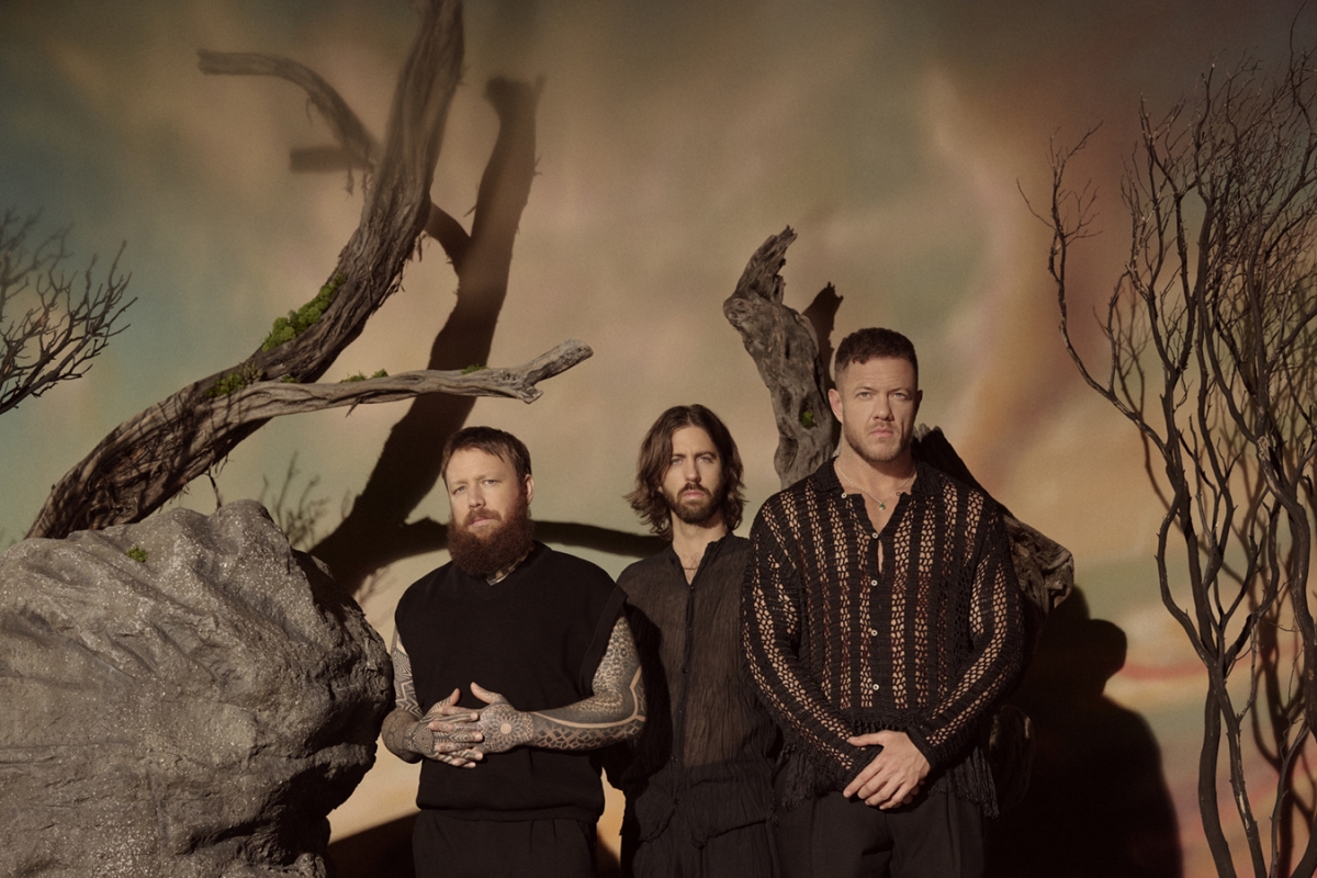 Three adult males pose indoors against a muted backdrop featuring bare tree branches and a textured gray boulder, creating a contemplative scene.