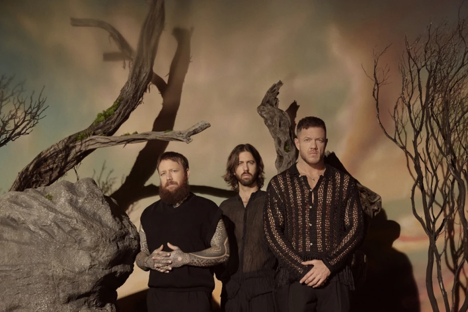 Three adult males pose indoors against a muted backdrop featuring bare tree branches and a textured gray boulder, creating a contemplative scene.