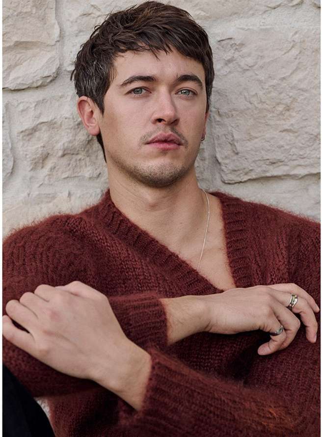 Portrait of an adult male with brown hair wearing a burgundy sweater, posing against a stone wall, looking directly at the camera.