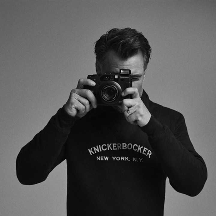 Monochrome portrait of a man holding a camera, wearing a black sweatshirt with 'Knickerbocker New York' text, against a grey background.