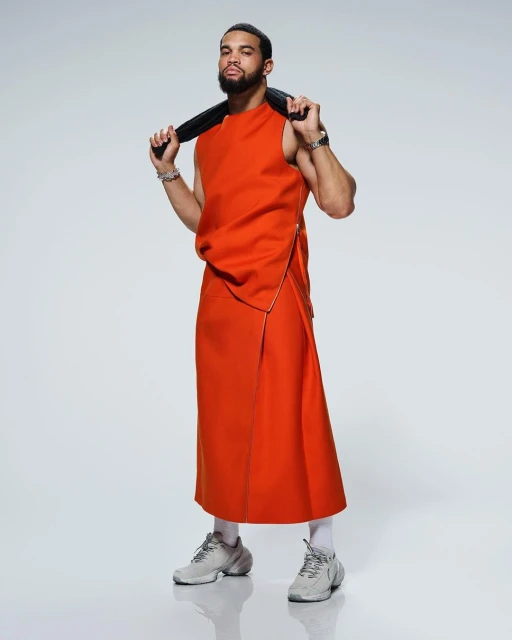 A confident Black adult male stands indoors in an orange sleeveless top and skirt, complemented by white sneakers, against a light backdrop.