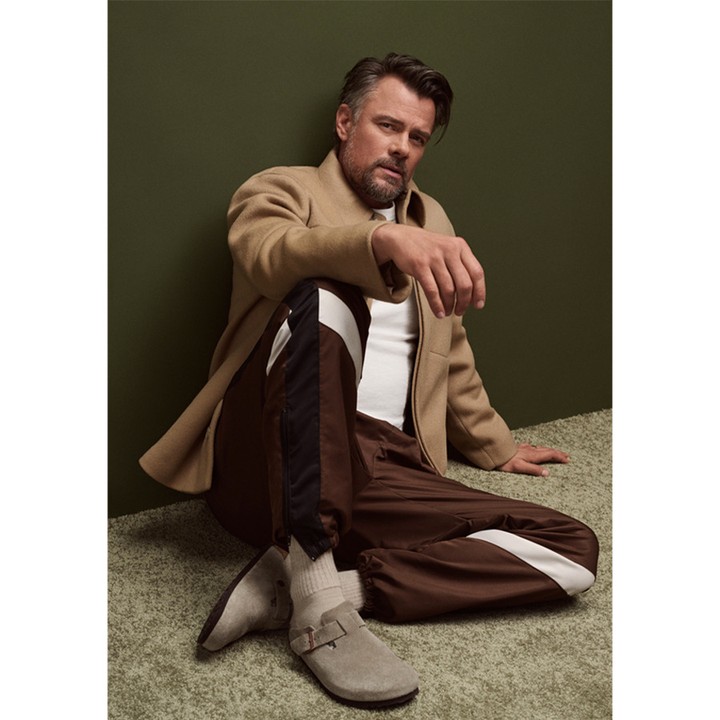 An adult male sits in a relaxed pose wearing casual apparel, including track pants, a t-shirt, a coat, and soft shoes with thick socks.
