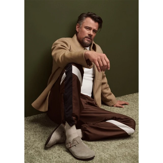 An adult male sits in a relaxed pose wearing casual apparel, including track pants, a t-shirt, a coat, and soft shoes with thick socks.