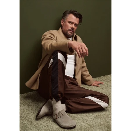 An adult male sits in a relaxed pose wearing casual apparel, including track pants, a t-shirt, a coat, and soft shoes with thick socks.