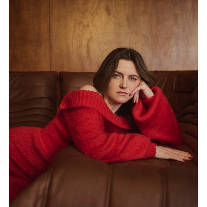 Brunette woman in red sweater dress poses on brown leather couch against a wood paneled wall in a portrait photograph indoors.
