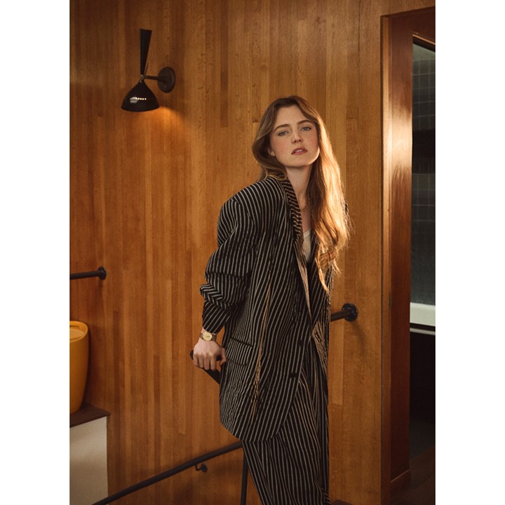 Woman with long brown hair poses in a striped pantsuit on wooden stairs next to a yellow vase and black sconce in a warm-toned interior.